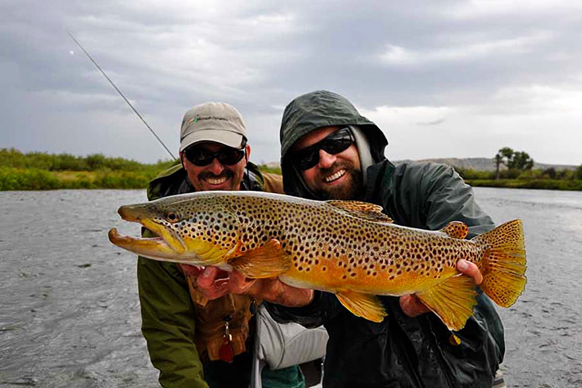 Two anglers holding a large spotted brown trout above a river